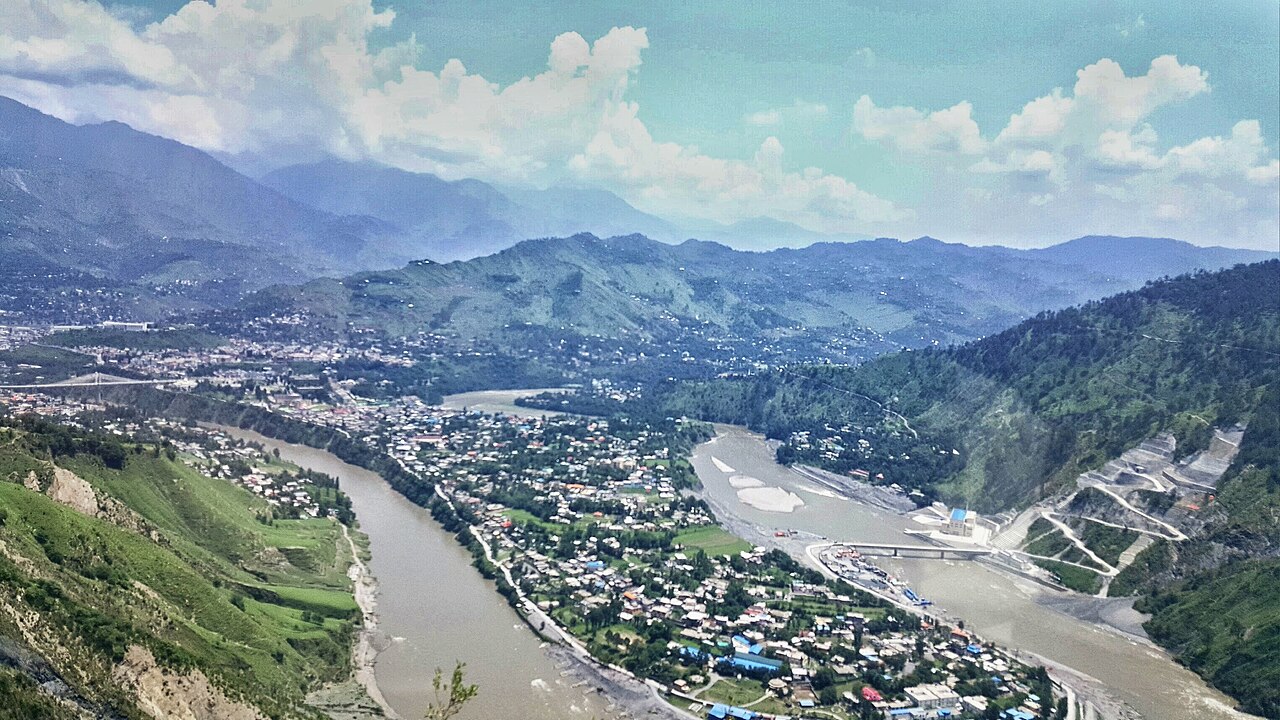 The Jhelum River winding through a green valley near Muzaffarabad, the capital city of Azad Kashmir, with forested hills rising in the background.