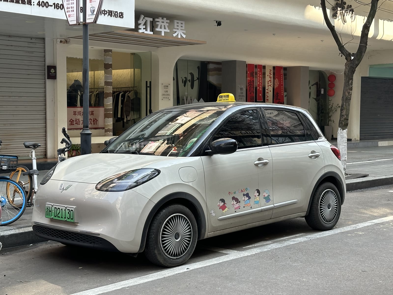 A pale grey Wuling Bingo electric hatchback parked on a city street beside a bicycle and a shopfront.