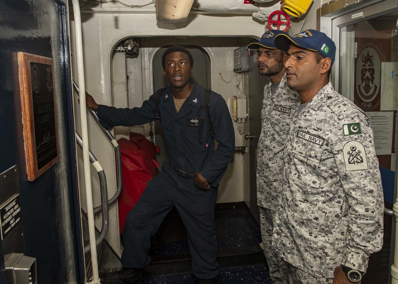 Two Pakistan Navy sailors in dark blue uniforms with peaked caps standing on the deck of a U.S. warship, looking at equipment on a console during a multinational naval exercise