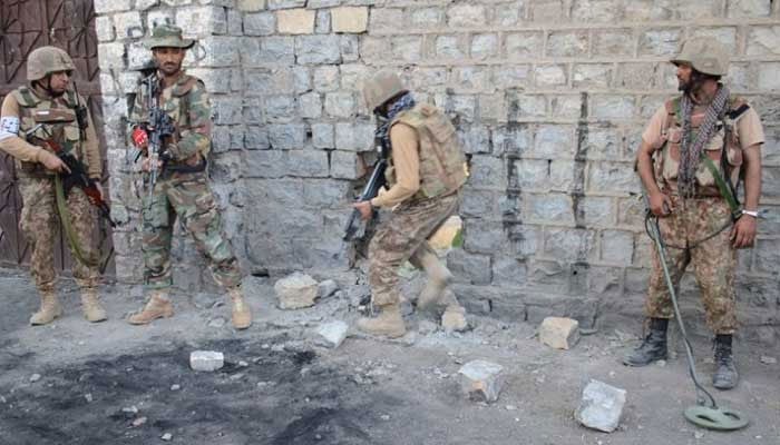 Soldiers in camouflage and helmets stand with rifles and metal detectors around a crater and rubble beside a stone wall during a security operation.