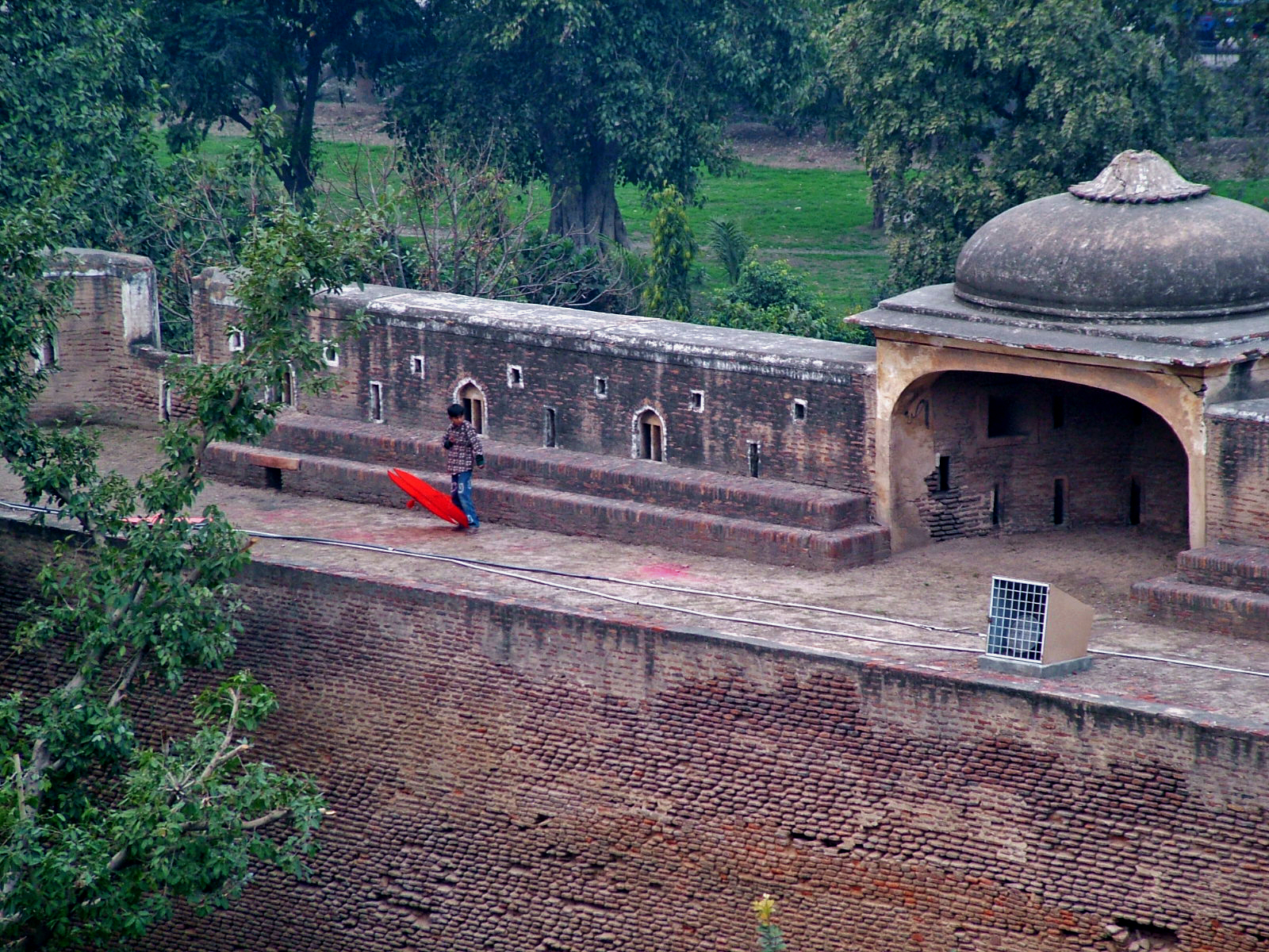 A boy crouches on a roof edge in Lahore preparing to fly a kite, with the red sandstone walls and towers of the Lahore Fort rising behind him against an overcast sky.