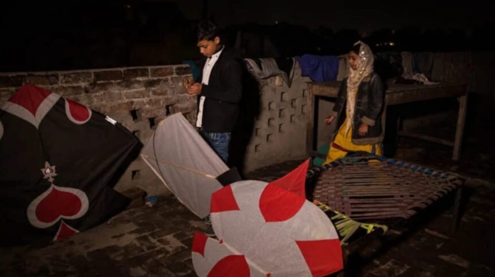 Two young people stand on a Lahore rooftop at night beside large kites bearing heart and spade playing-card designs.