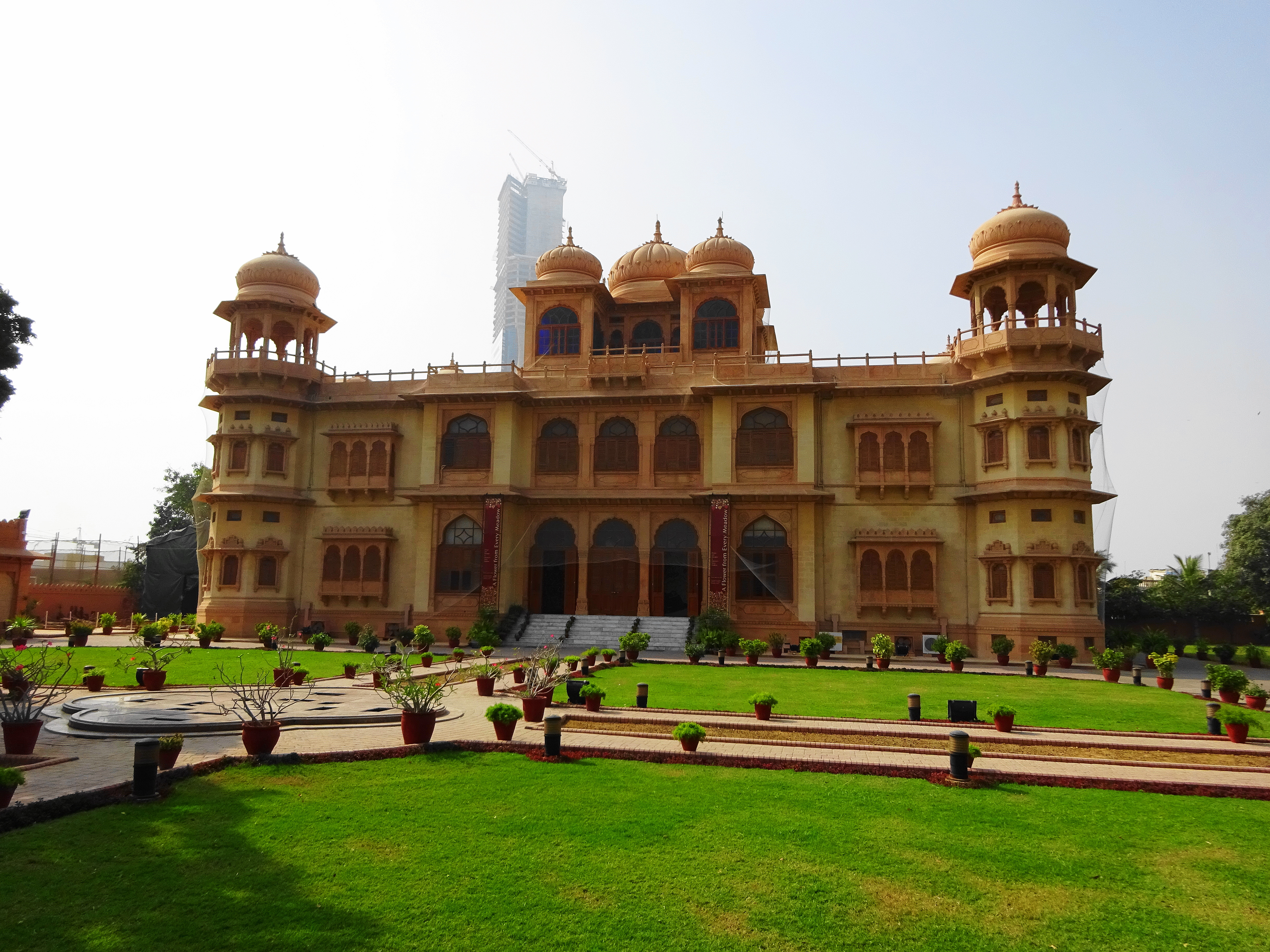 The yellow-stone Mohatta Palace Museum in Clifton, Karachi, photographed in daylight, showing its central dome, arched windows and surrounding trees against a clear sky.