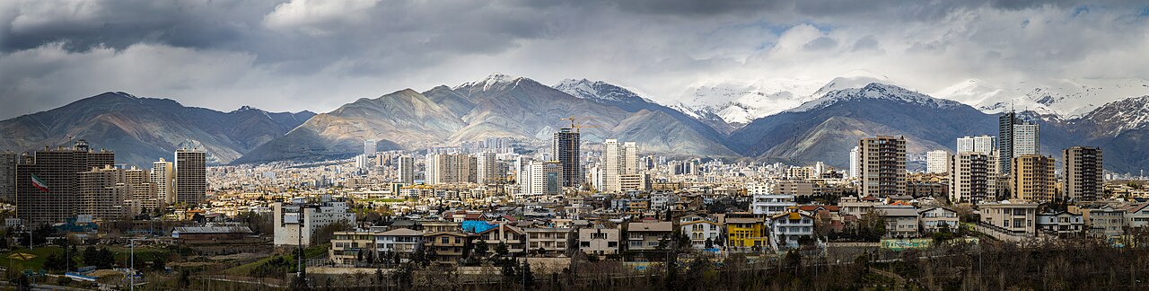 A wide panoramic view of northern Tehran against snow-capped mountains under a clear sky, with low-rise city buildings stretching across the foreground.