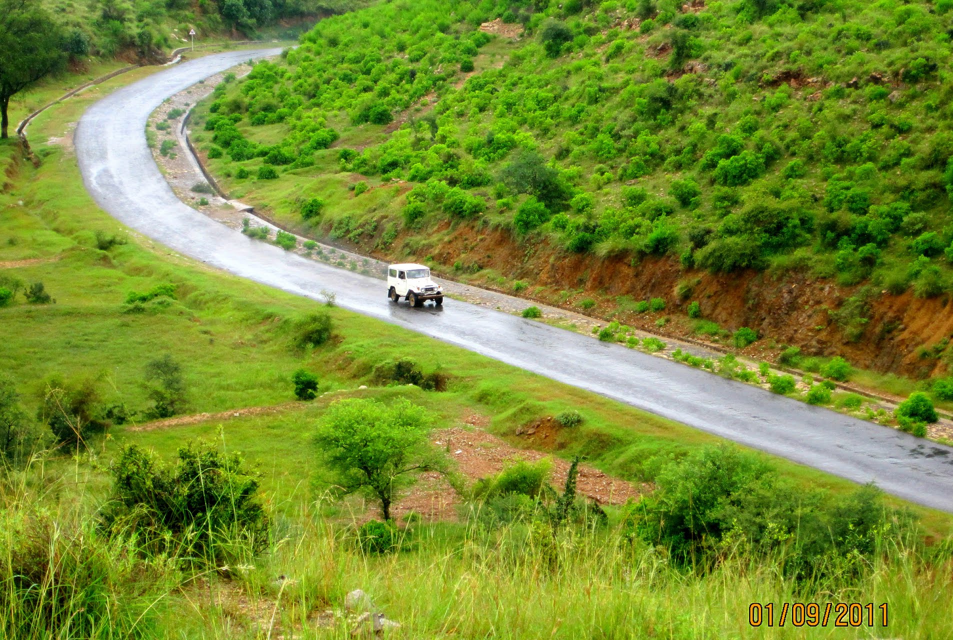 A winding two-lane road curving through fog-covered green hills in the Salt Range of Punjab, Pakistan.