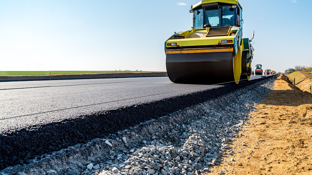 A yellow road roller compacting fresh black asphalt on a new road surface beside a gravel shoulder.