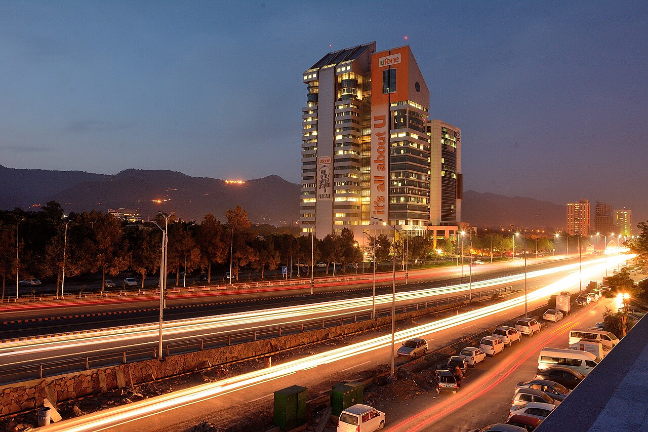 A long-exposure night view of the Blue Area commercial district in Islamabad, with lit office buildings and light trails along a central avenue.