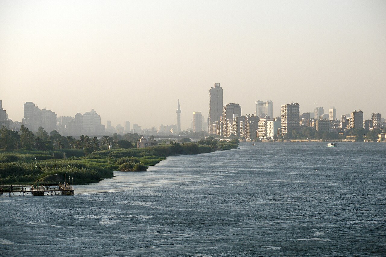 A wide daytime view of the Cairo skyline along the Nile, with low-rise buildings stretching into the distance and the green river and a bridge visible in the foreground under a hazy sky.