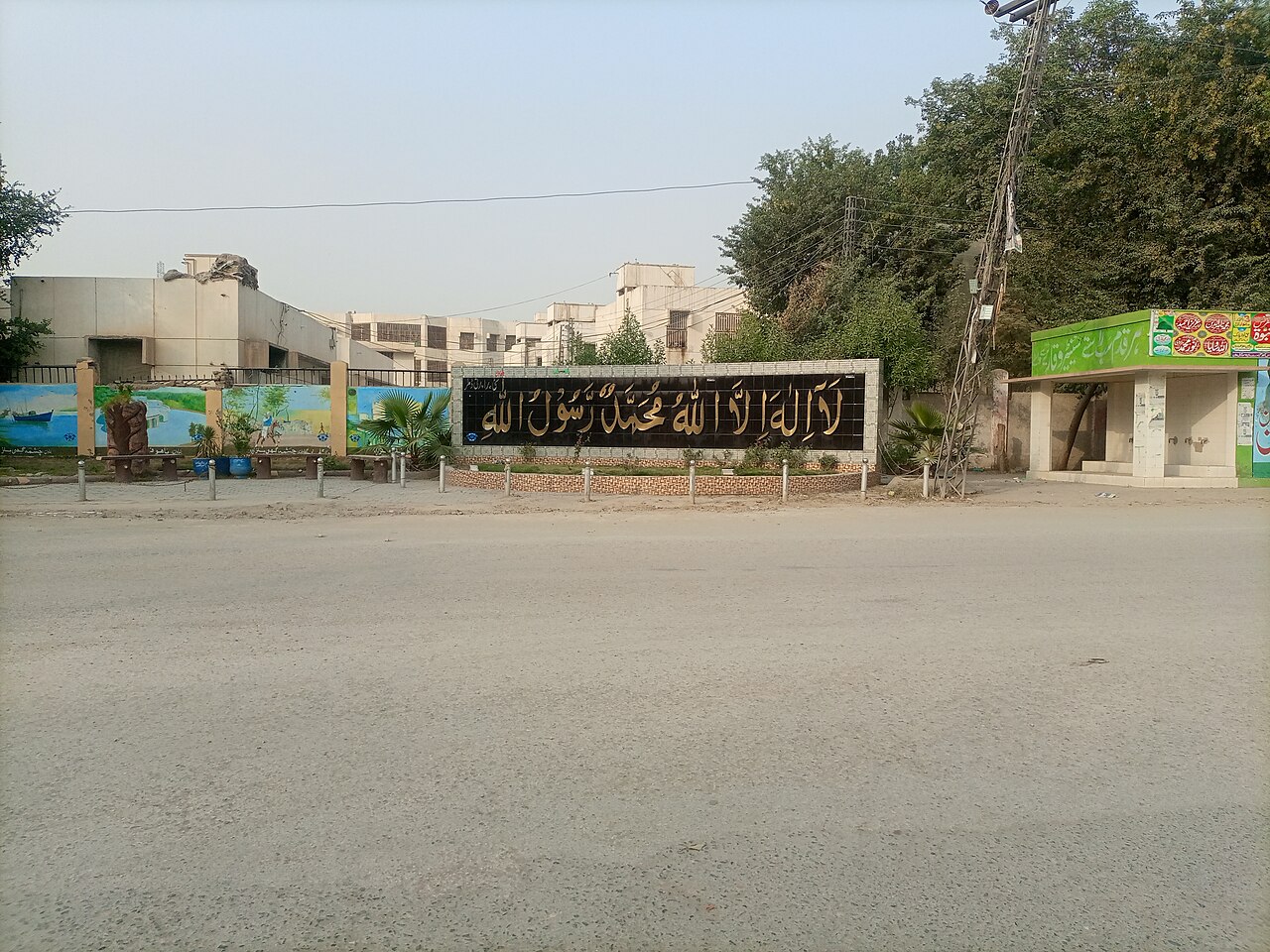 A wide, empty road and roundabout in Dera Ismail Khan, with a large black calligraphy wall and low pale buildings behind a line of trees.