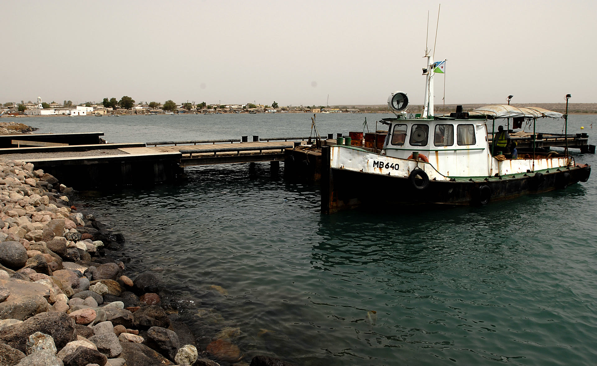 A small tugboat moored beside a low concrete pier at the port of Obock in Djibouti, with calm water and hazy hills behind.