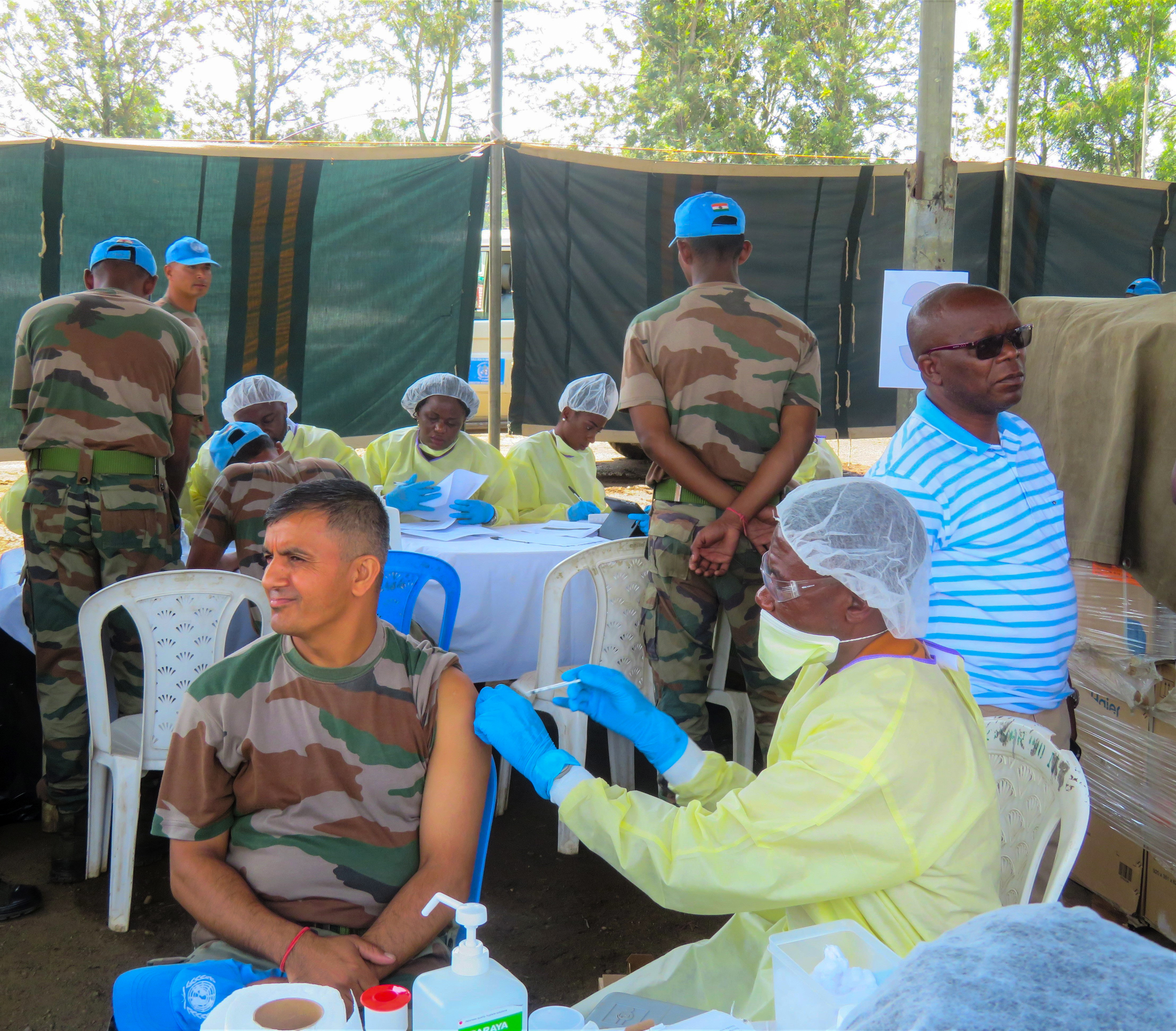 A health worker in a yellow protective gown vaccinates a seated UN peacekeeper outdoors in eastern Congo