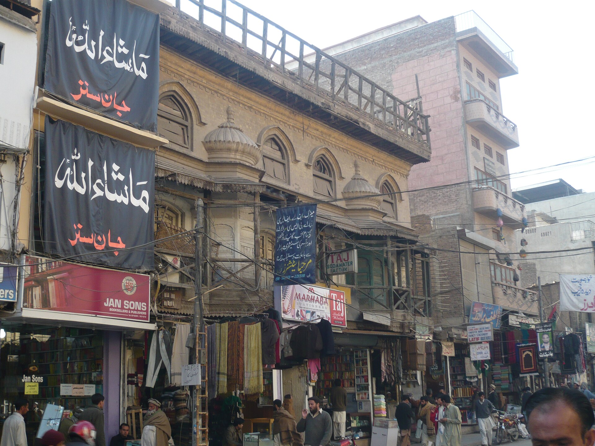 A busy market street in Peshawar's old city, with Urdu and English shop signs, bolts of cloth hanging outside the stalls, motorbikes at the kerb and pedestrians passing an old building with arched windows and domed turrets.