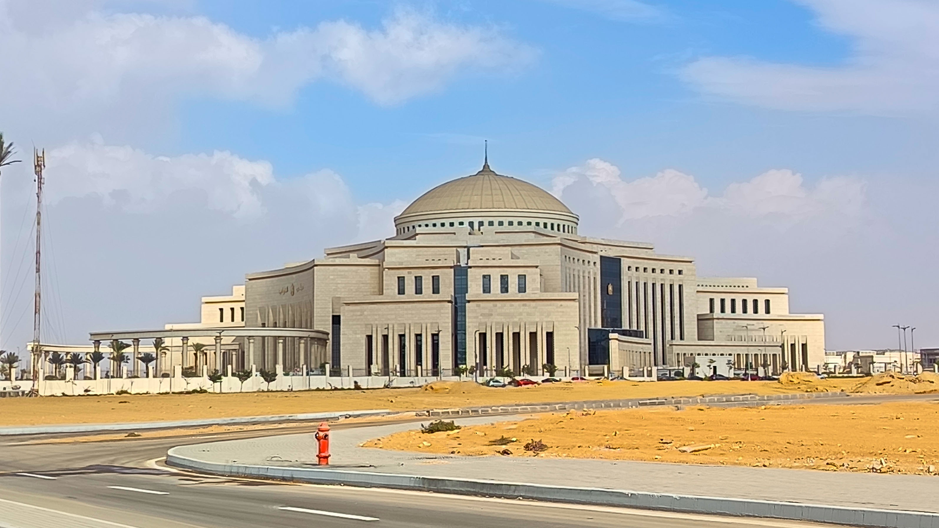 The domed stone facade of Egypt's parliament building in the New Administrative Capital, seen across an empty road and sandy forecourt under a blue sky.