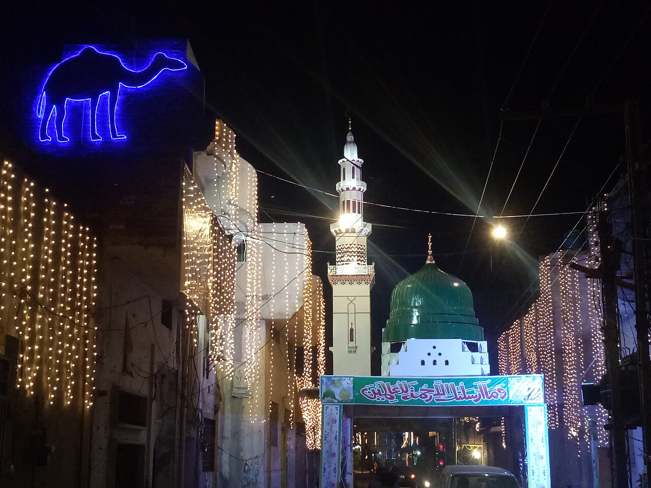 A street at night decorated with strings of warm lights beside a floodlit mosque with a green dome, marking Eid Miladun Nabi in Pakistan.