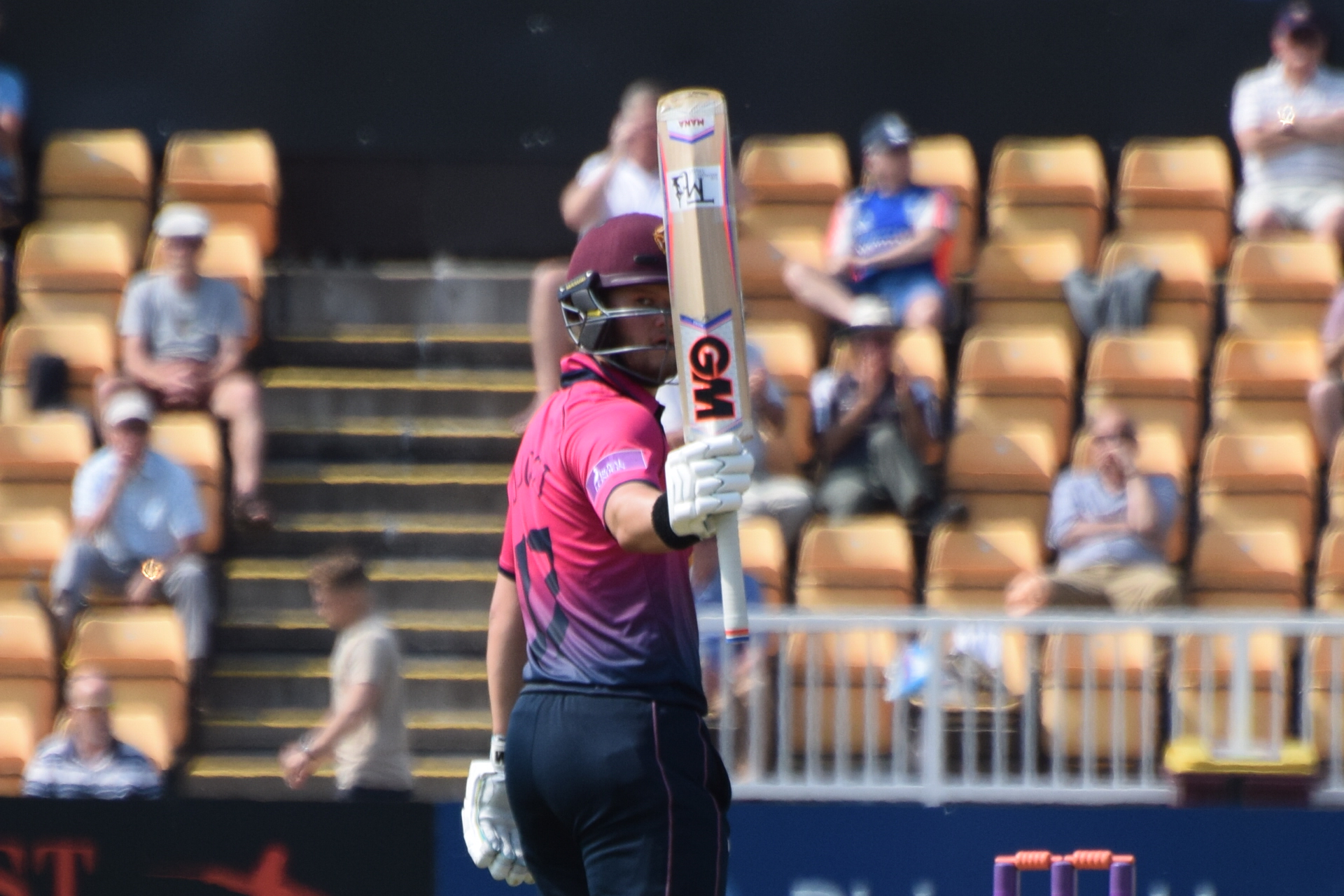 A batter in a pink and maroon shirt and maroon helmet raising his bat upright in front of his face, with blurred spectators in tan seats behind him.