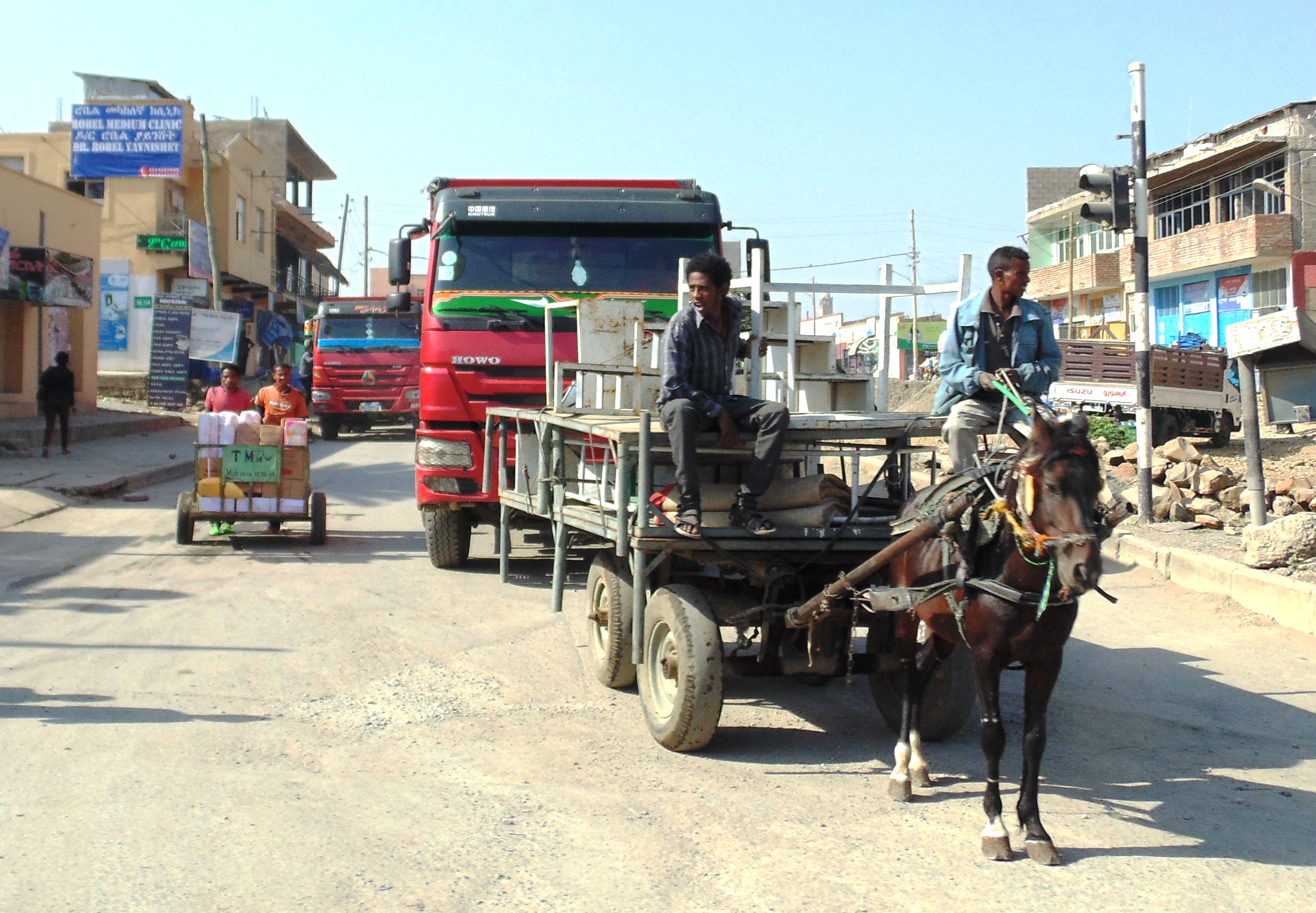 A street in Mekelle, Ethiopia, with a horse-drawn wooden cart in the foreground, a red lorry, a handcart and low shop buildings along a wide road under a clear blue sky.