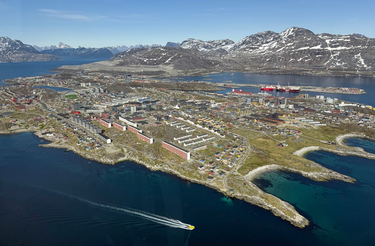 An aerial view of a coastal town of colourful low-rise buildings beside deep blue water, with snow-streaked mountains behind and a small boat trailing a white wake.