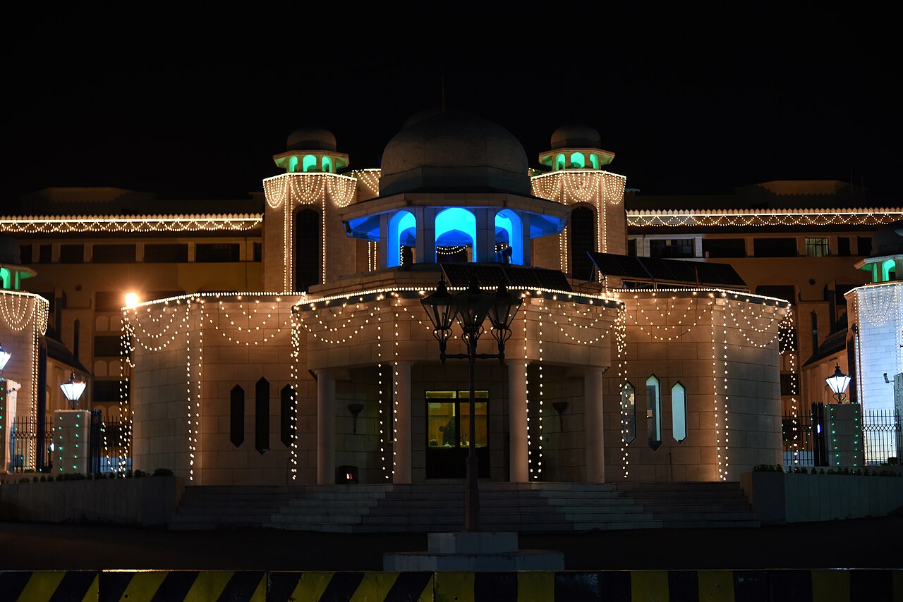 A floodlit government building in Islamabad at night, with a wide paved forecourt, rows of windows and a low central entrance.