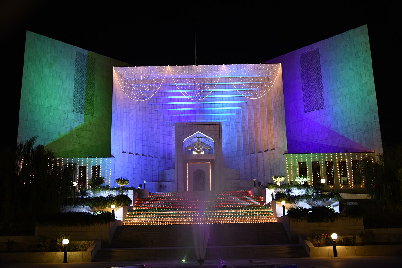 The Supreme Court of Pakistan building in Islamabad, photographed at night from the air, its tiered roofline lit against the dark sky and the surrounding gardens and access roads in shadow.