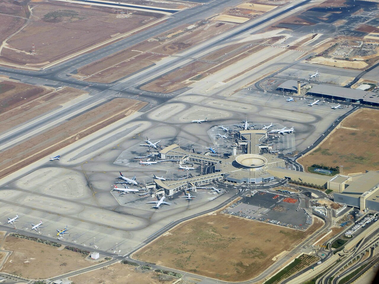 An aerial view of Ben Gurion Airport, showing its terminal buildings, aprons, control tower and runways set in flat, built-up terrain.