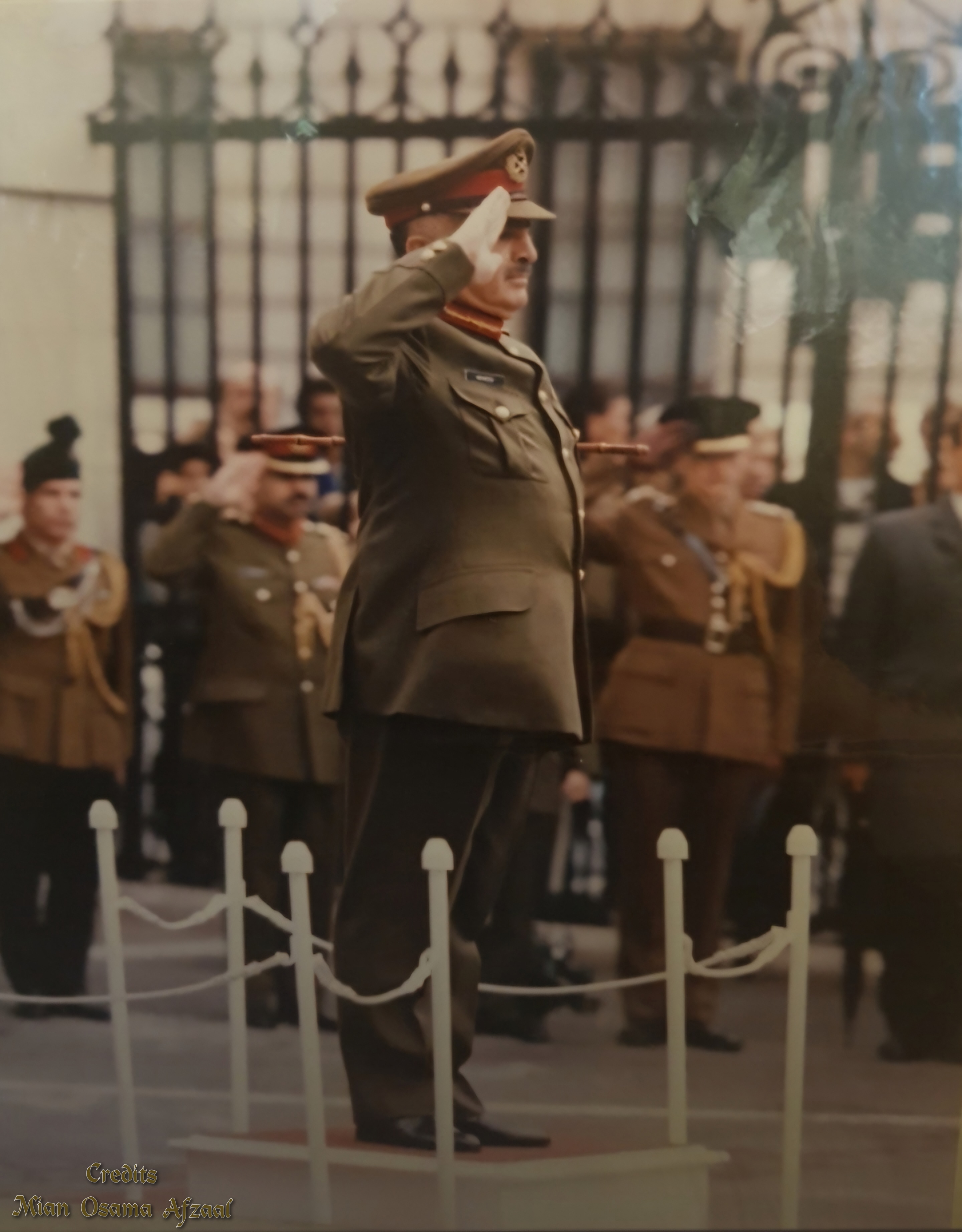 General Abdul Waheed Kakar in army uniform and peaked cap, saluting with a white-gloved hand at a ceremonial parade, with ranks of soldiers and a white picket fence behind him.