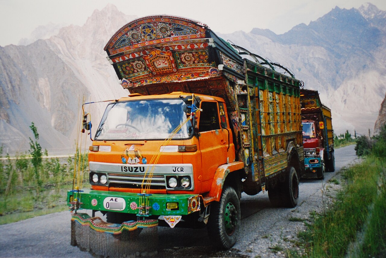 A brightly painted orange truck with a decorated wooden crown travels along a gravel mountain road, with another truck behind it.