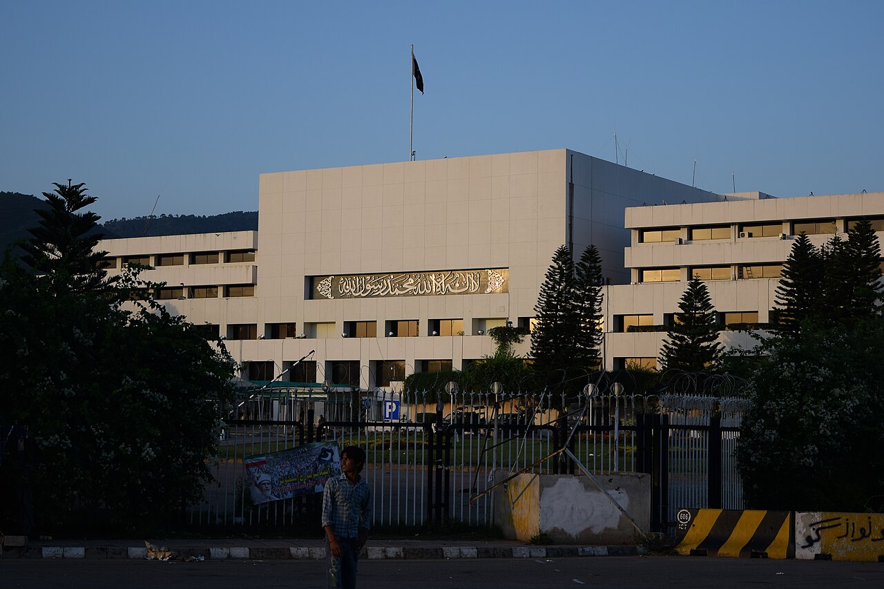 The Pakistan Parliament House in Islamabad at dusk, a modern multi-storey block with the Bismillah calligraphy panel above the entrance and the Pakistani flag flying on the roof, with a boy standing in front of the gated perimeter