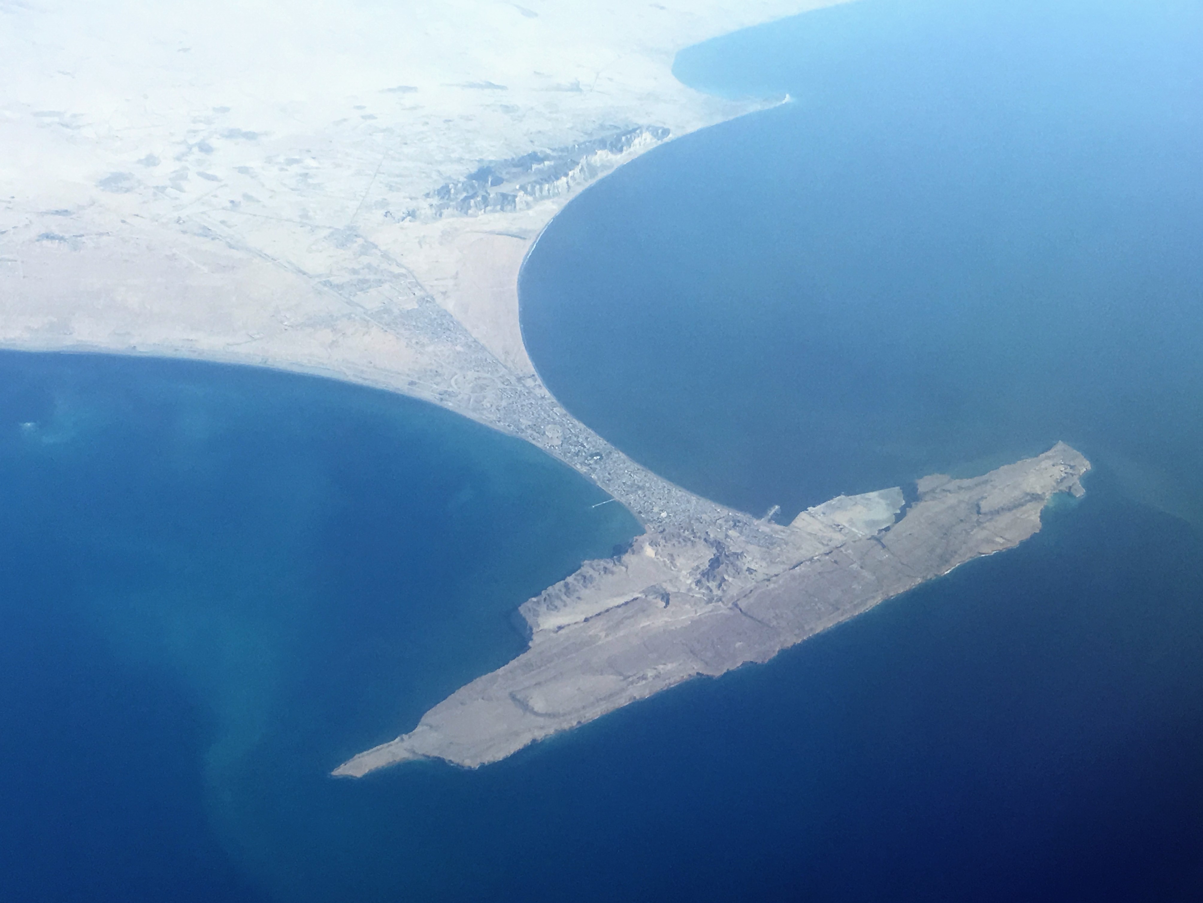 An aerial view of a coastal city on a narrow peninsula beside deep blue sea, with bare tan mountains and a crescent-shaped bay behind the built-up area.
