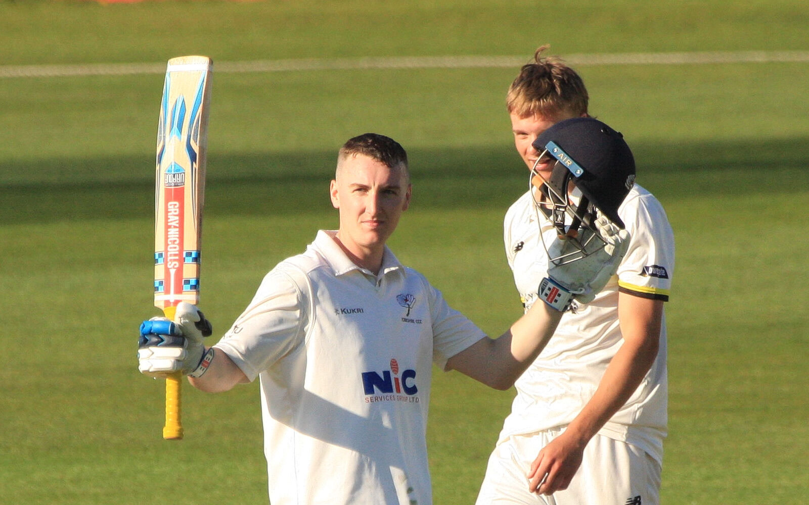 Harry Brook in white Yorkshire kit raising his bat at the crease, with a team-mate holding a helmet beside him