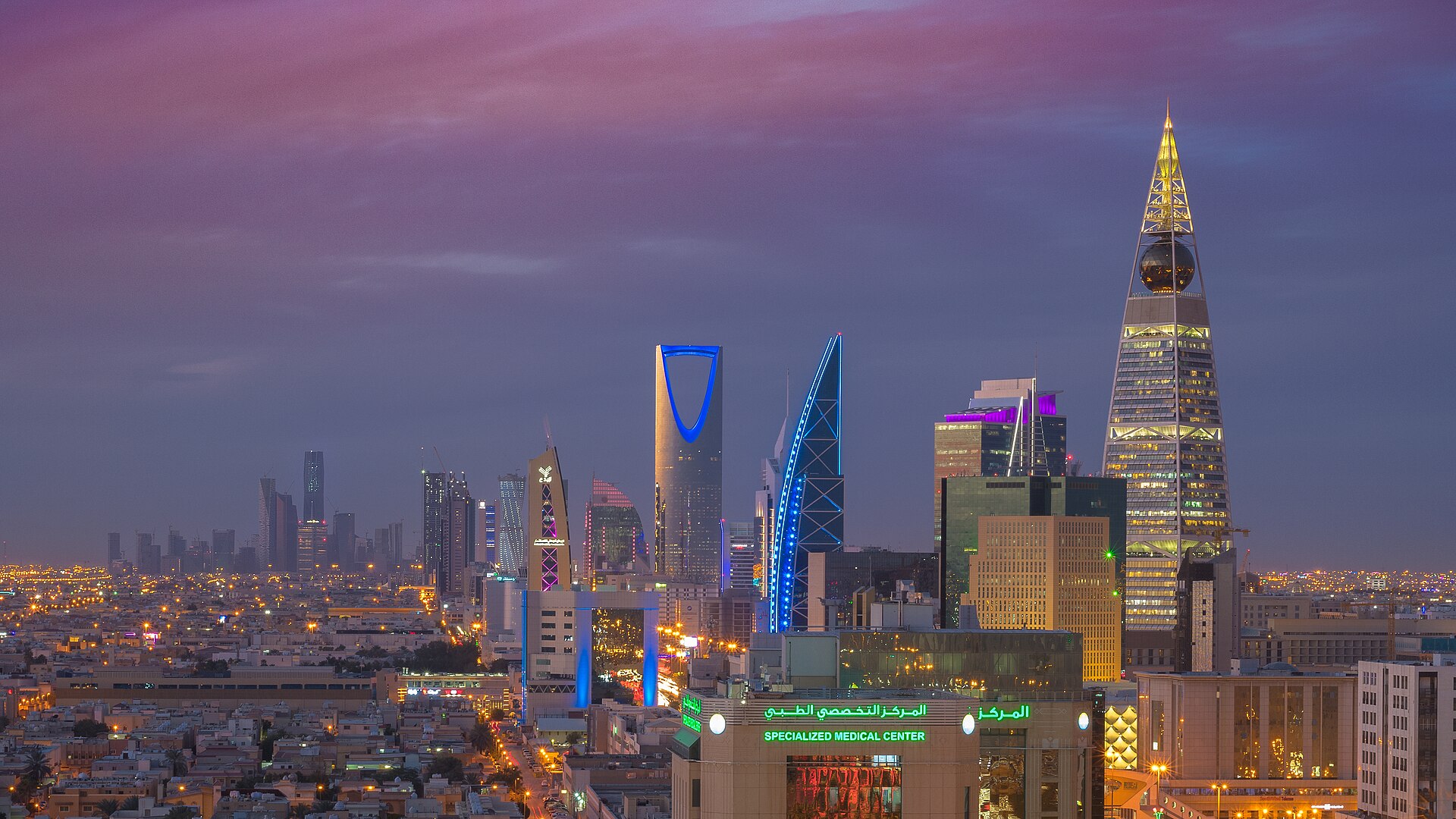 Riyadh's city skyline at night, with the illuminated Kingdom Centre tower and other high-rise buildings rising over lit roads and trees.