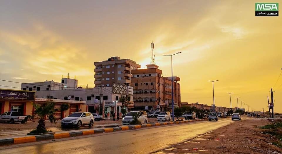 A street in Ataq, the capital of Shabwa governorate, at sunset, with cars along the curb, low commercial buildings, a water tower and streetlights beneath an orange sky.