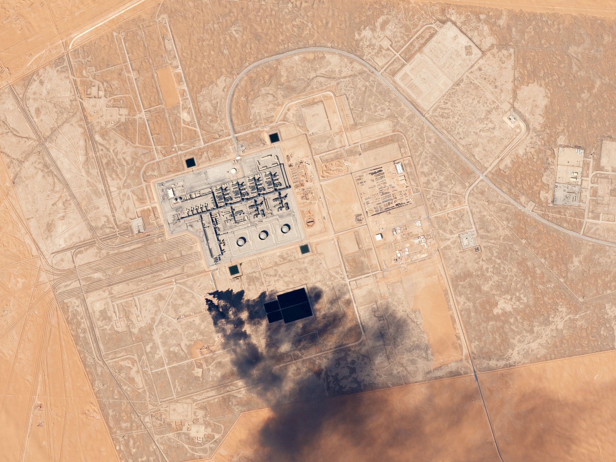 A satellite image of Saudi Aramco's Khurais oil processing facility, showing industrial plant structures, storage tanks and access roads in desert terrain.
