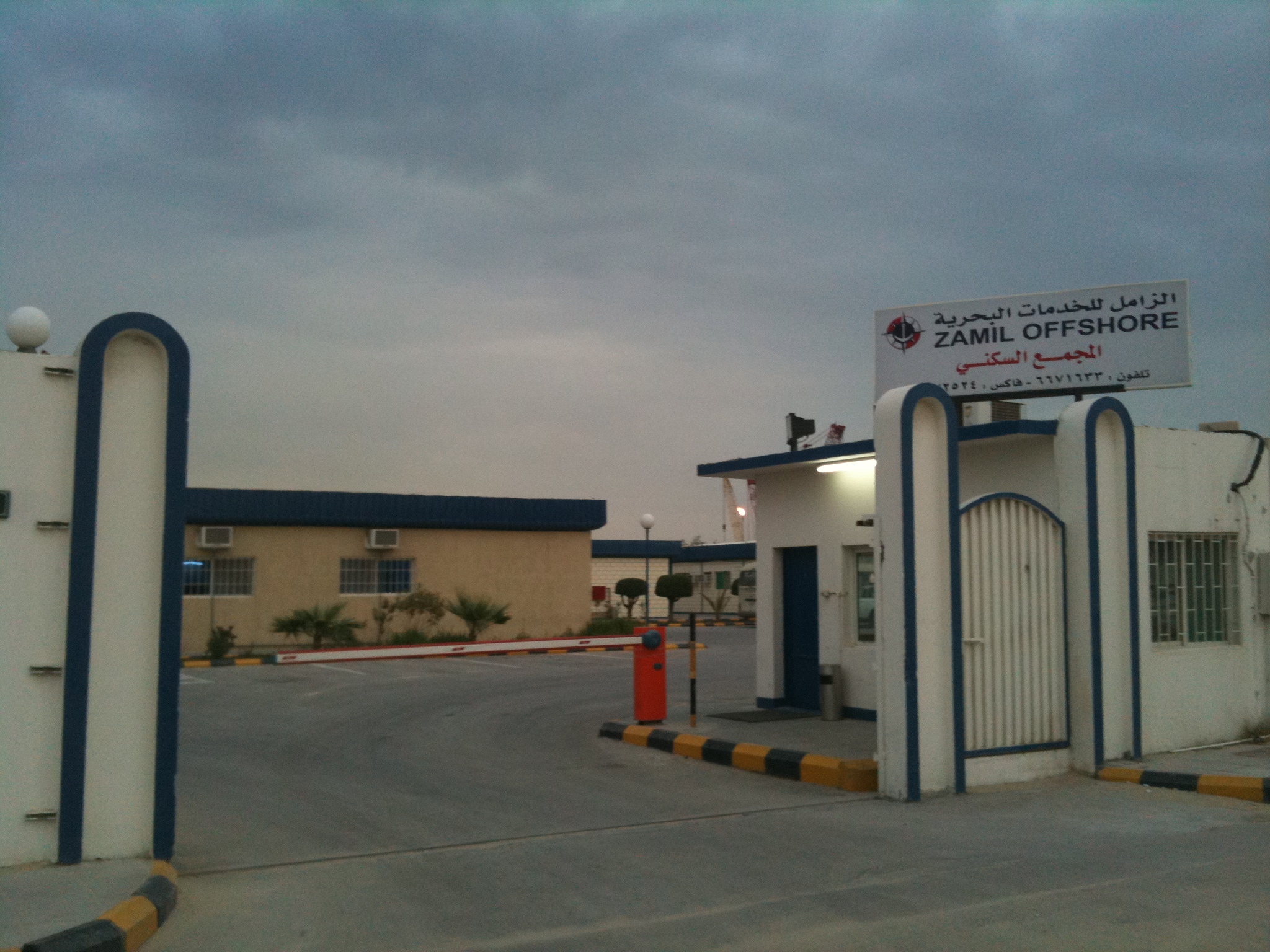 The entrance gate of the Zamil Offshore compound at Ras Tanura in Saudi Arabia's Eastern Province, with a bilingual Zamil Offshore sign in English and Arabic above a red and white barrier arm and a guardhouse under an overcast sky.
