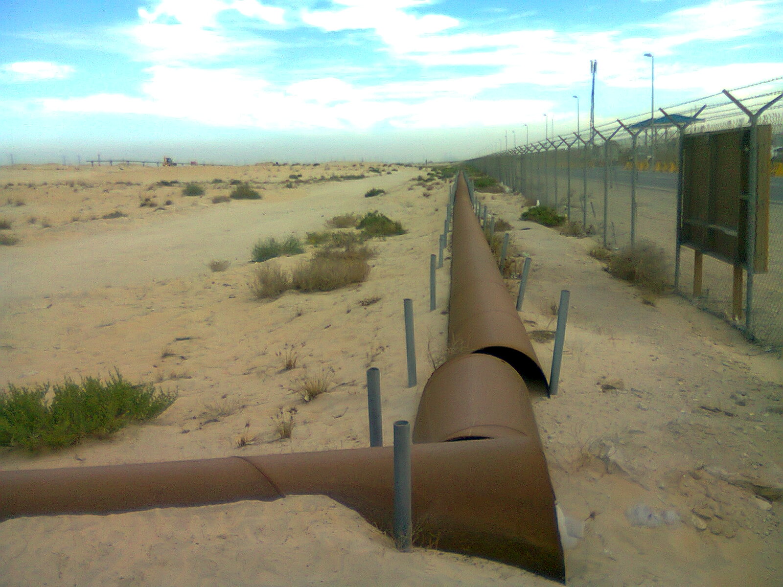 A large rust-coloured above-ground oil pipeline running across sandy desert beside a barbed-wire security fence, with sparse shrubs and distant vehicles on the horizon.