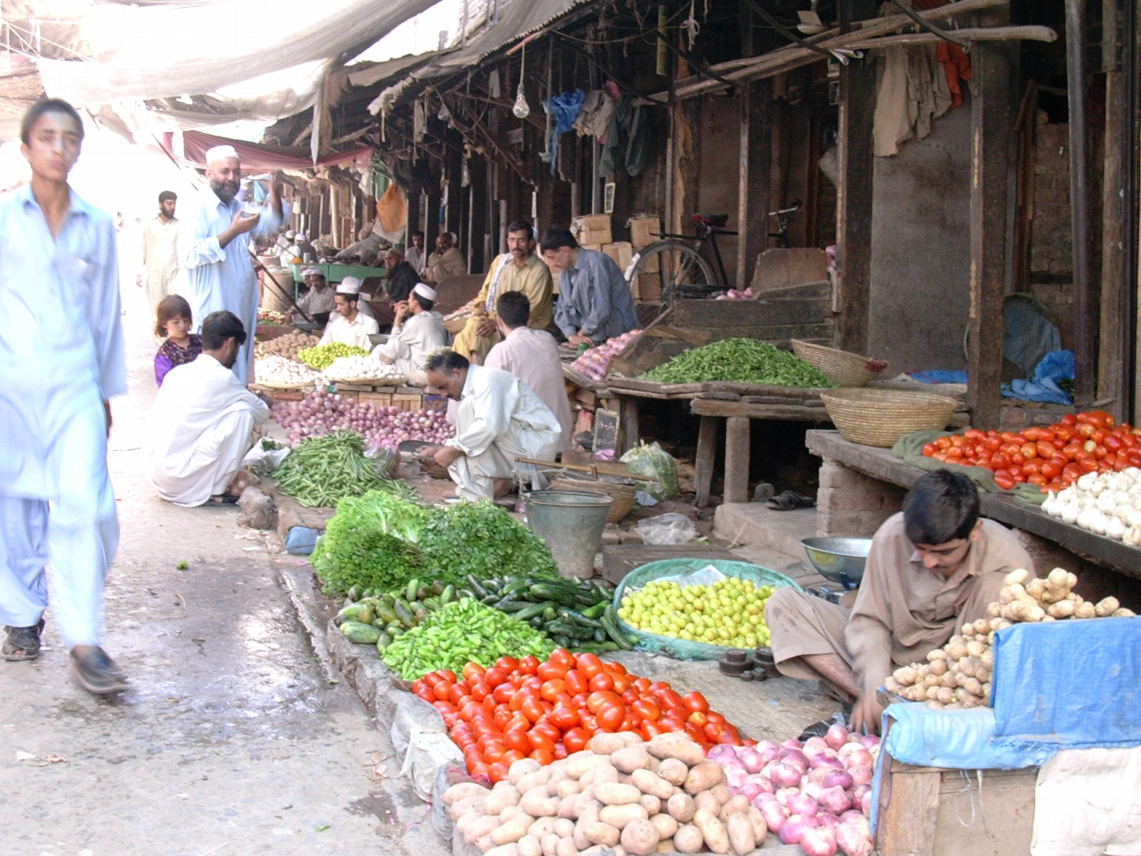 A busy open-air vegetable market in Peshawar, with vendors seated behind wide baskets of produce and shoppers moving between the stalls.