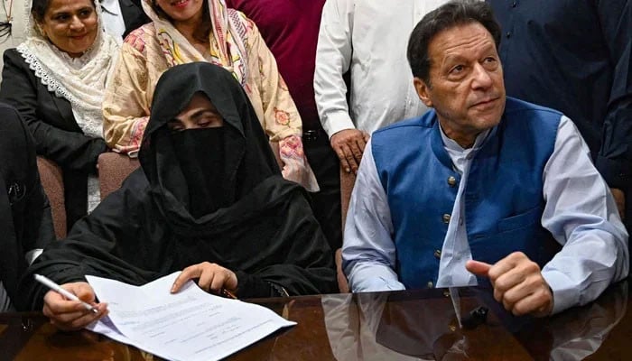 Imran Khan, wearing a blue waistcoat over a light shirt, seated at a table beside Bushra Bibi, who is in a black niqab and signing a document.