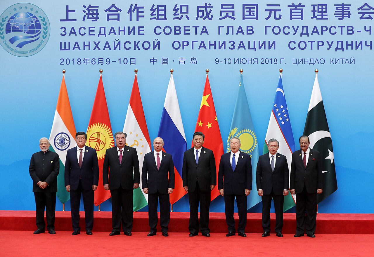 Heads of state of the Shanghai Cooperation Organisation member states posing for a family photograph at the 2018 Qingdao SCO summit, framed by their national flags.