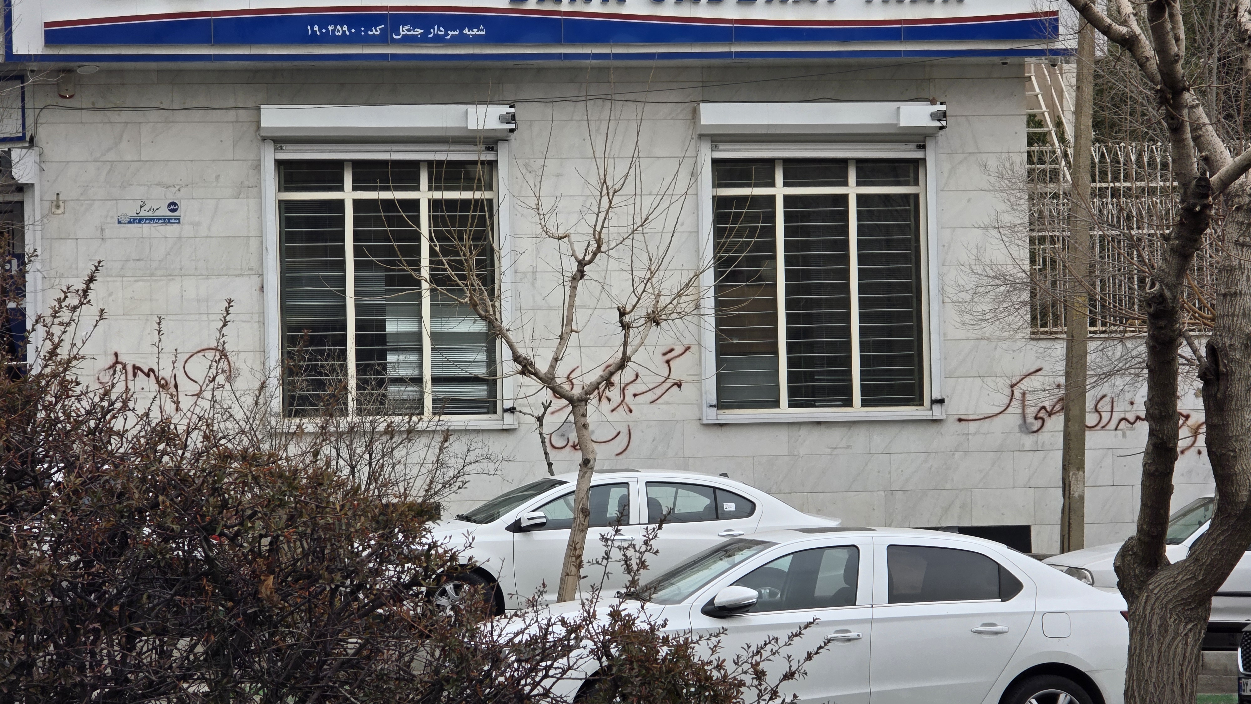 A Bank Saderat Iran branch in Tehran with Persian-script protest graffiti on the marble wall outside, photographed on Sardar-e Jangal Boulevard