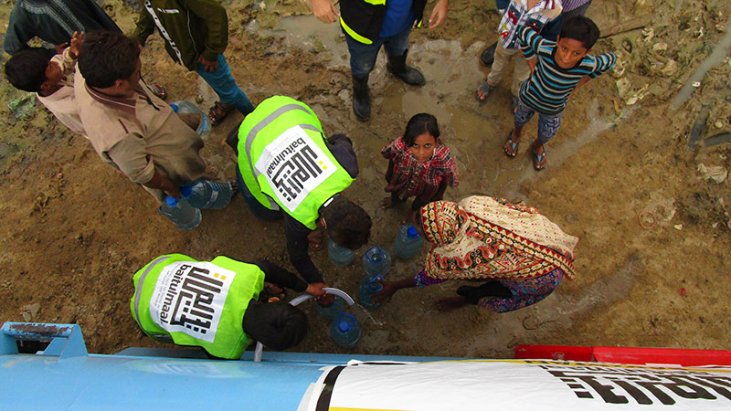 An overhead view of rescue workers in hi-vis vests and residents standing in muddy floodwater beside a rescue boat, with children and water containers nearby.