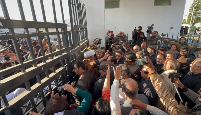 A crowd of opposition supporters and media gathered behind a black metal gate outside a white building