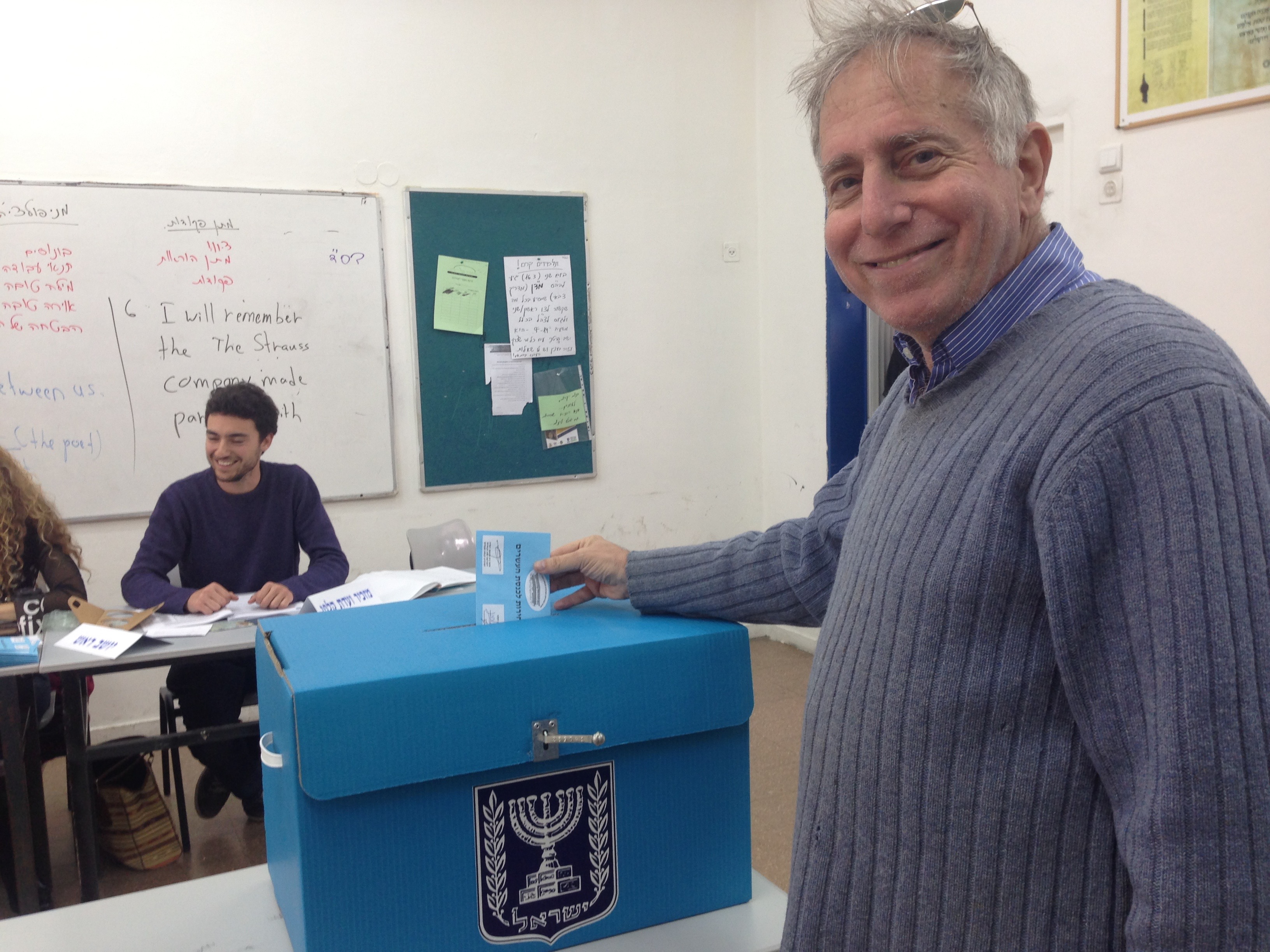 A voter holds a blue ballot envelope over a blue ballot box printed with Israel's national emblem and the Hebrew word for elections, inside a polling station where officials sit at a table beside whiteboards covered in handwritten campaign notes.