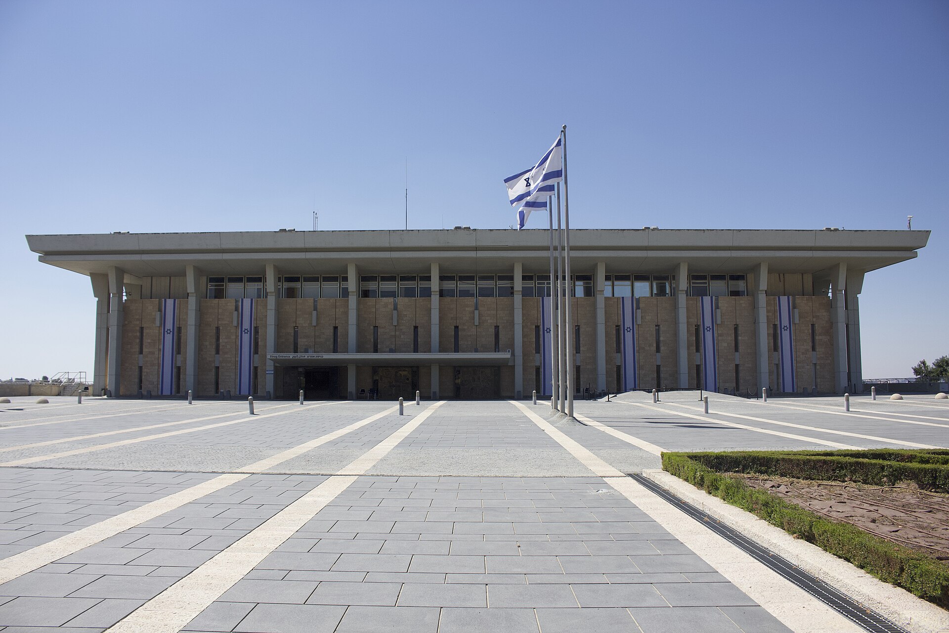 The wide stone and concrete facade of the Knesset in Jerusalem, hung with vertical blue-and-white banners bearing the Star of David, with an Israeli flag flying on a tall pole in the paved plaza in front.