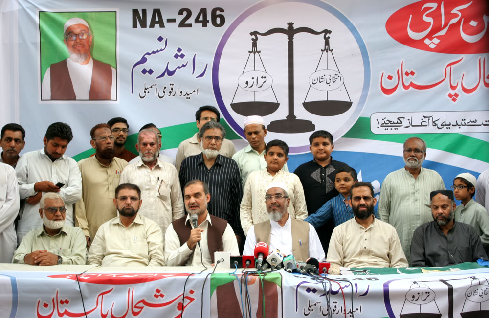 Jamaat-e-Islami supporters sit behind a table of microphones beneath a green-and-white party banner at an outdoor political gathering.
