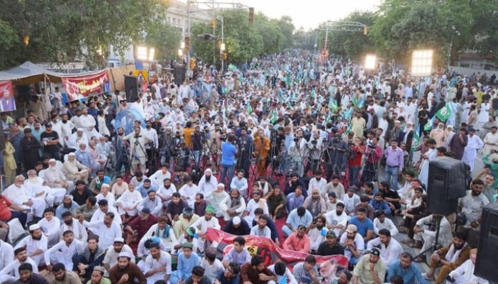 A large crowd of men seated and standing at an outdoor political gathering on a city street at dusk, with green flags and a stage.