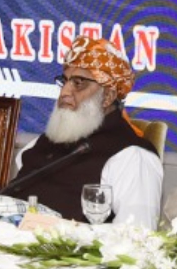 Maulana Fazlur Rehman, wearing an orange patterned turban, glasses and a white beard, sits in a dark waistcoat in front of a banner reading Pakistan.