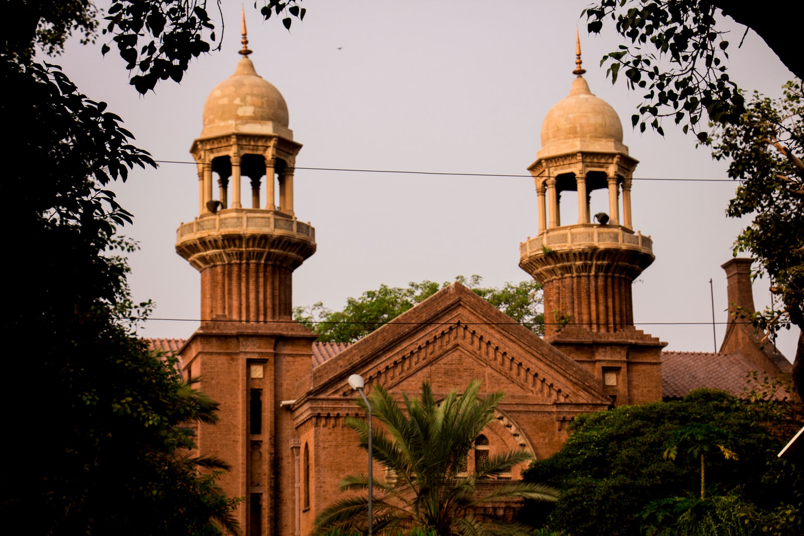 The historic red-brick Lahore High Court building on the Mall Road in Lahore, with two domed corner towers above an arched entrance, framed by trees.