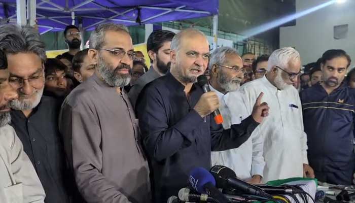 Jamaat-e-Islami leader Hafiz Naeemur Rehman speaks into a microphone at an outdoor press conference, flanked by other party leaders and a cluster of media microphones.