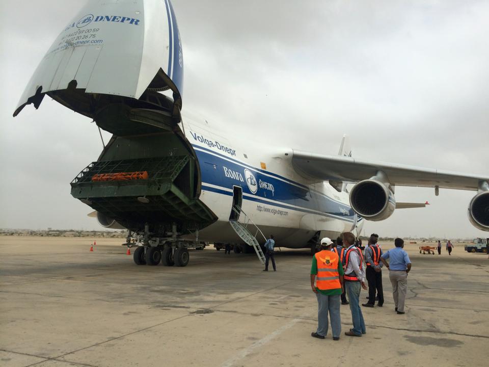 A Volga-Dnepr Antonov An-124 cargo aircraft on the apron at Jinnah International Airport with its rear cargo door raised and a loader positioned underneath, while ground staff in orange high-visibility vests stand nearby.