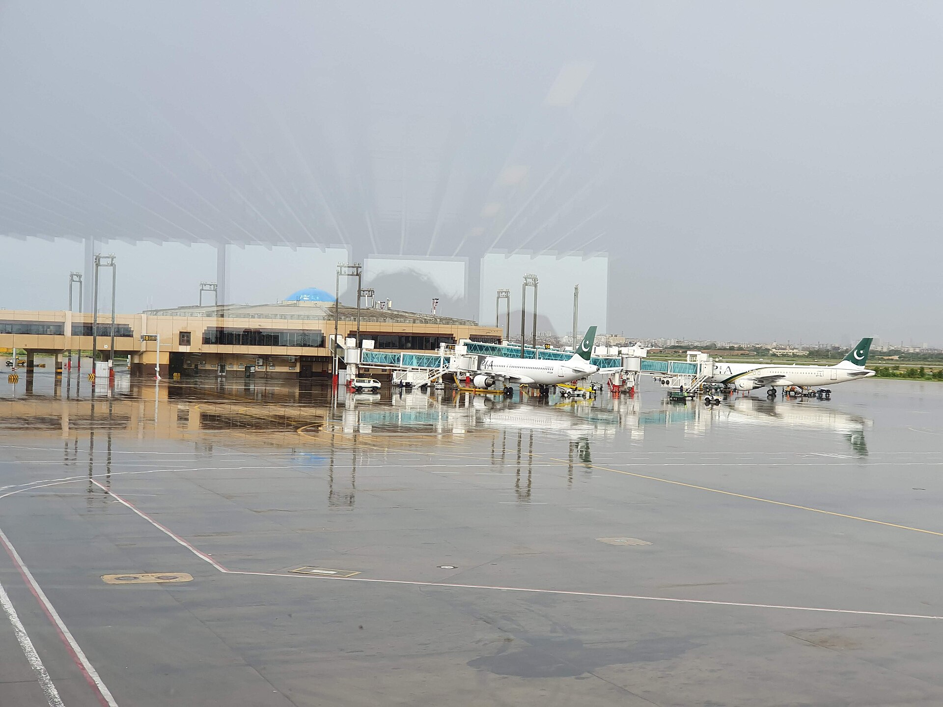 Wide view across the wet apron at Jinnah International Airport in Karachi, with two Pakistan International Airlines Boeing 737s parked at gates linked to jet bridges, the terminal building behind them and a hazy city skyline on the horizon.