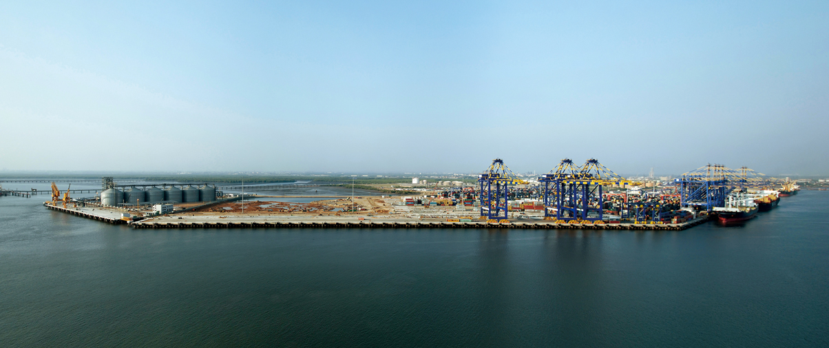 Aerial view of a container port with blue gantry cranes, stacked shipping containers, a berthed vessel and oil storage tanks